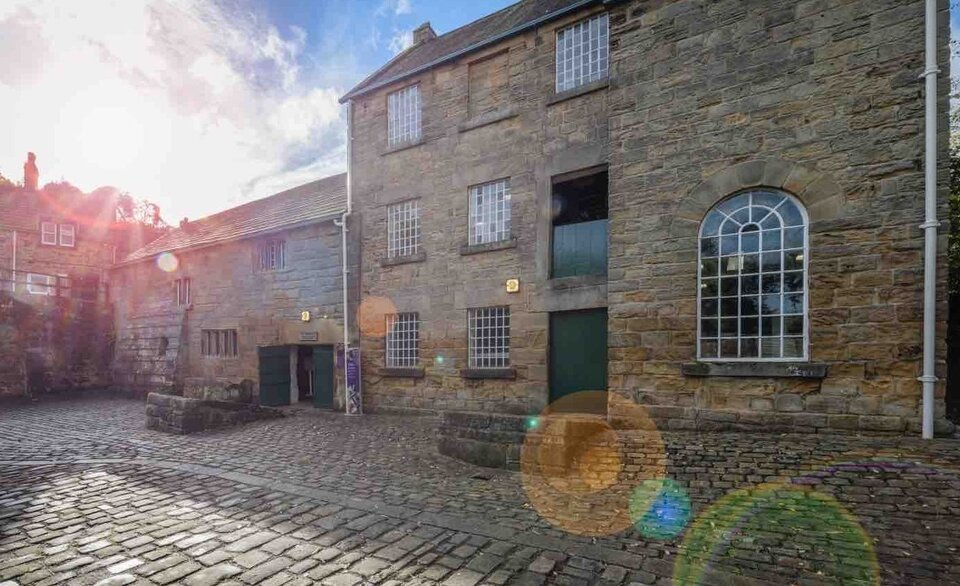 An old stone building with multiple windows and doors, set in a cobblestone courtyard under bright sunlight with lens flares in the foreground.